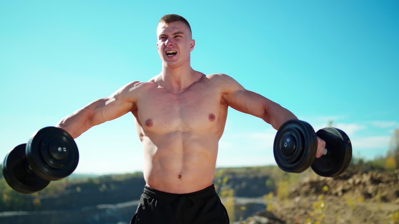 Portrait of concentrated sportsman lifts dumbbells. Strong bodybuilder training with heavy dumbbells on the nature background in summer.