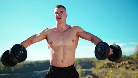 portrait of concentrated sportsman lifts dumbbells. strong bodybuilder training with heavy dumbbells on the nature background in summer.