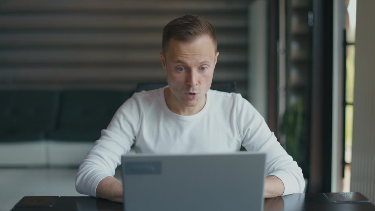 Happy man sits in room at desktop with laptop and rejoices in his success. Man actively gesticulates with his hands in delight. Slow motion