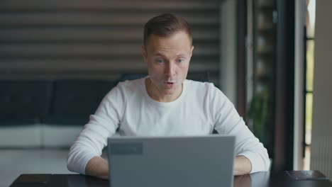 happy man sits in room at desktop with laptop and rejoices in his success. man actively gesticulates with his hands in delight. slow motion