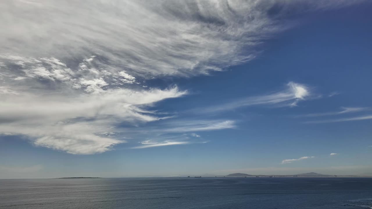 Wispy white clouds in a blue sky over a calm ocean, with Robben Island visible in the distance, and neutral space for text titles.