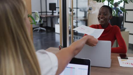 caucasian woman giving a document to her african american female colleague at office