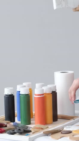 close-up of a hand with various art supplies, including colorful paint bottles, brushes, and a palette on a table