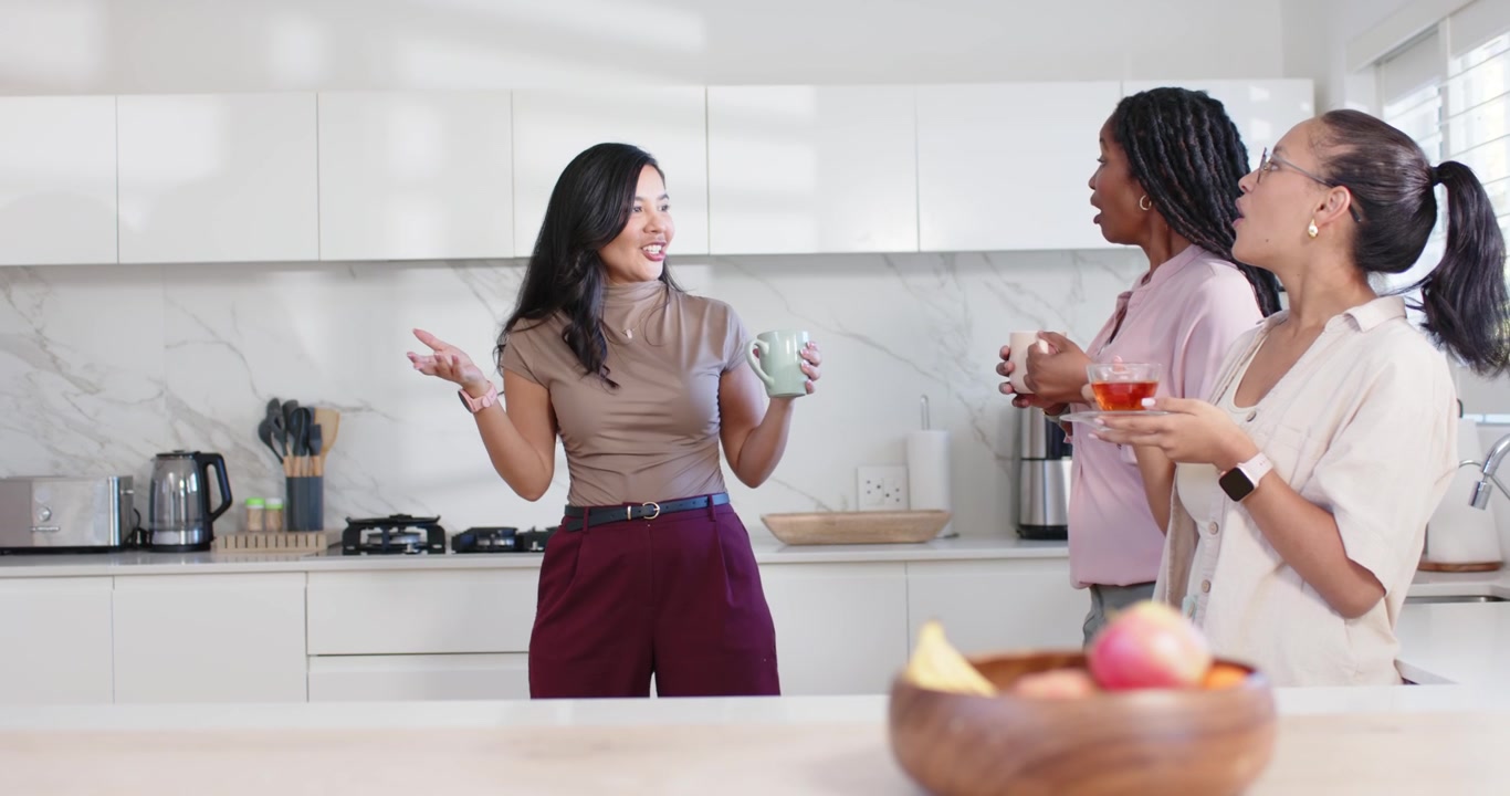 Amiche diverse con tazze in una cucina luminosa, la donna al centro che si muove e racconta una storia.