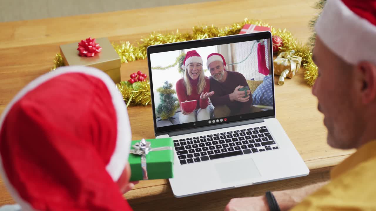 padre y hijo caucásicos con sombreros de santa usando una computadora portátil para una videollamada de navidad con una pareja en la pantalla