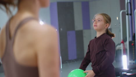 close-up of woman in brown and kid in glasses juggling, tossing green balloon into air, mirror reflecting another child in purple top working out in modern gym