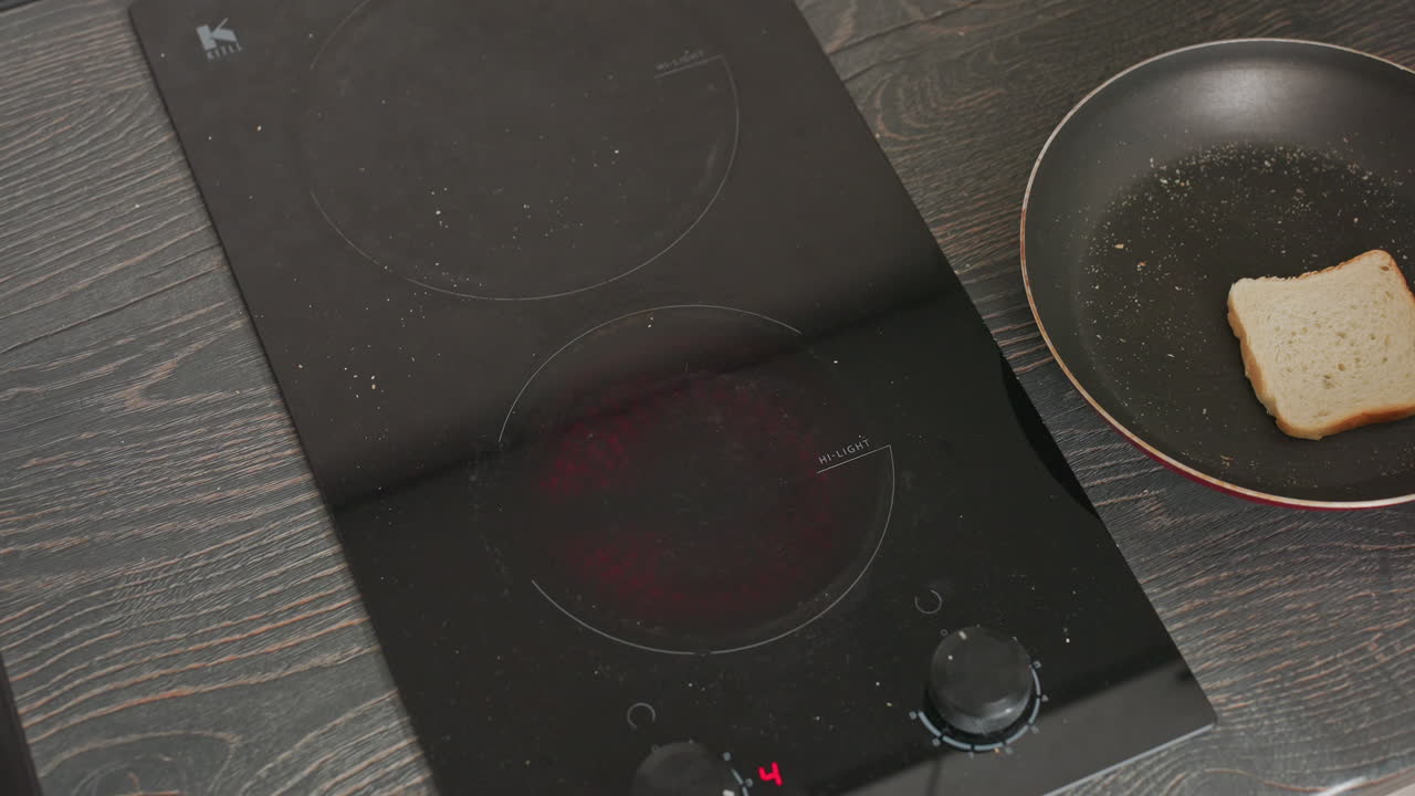 Vista dall'alto di una persona dalla pelle chiara che regola una manopola per accendere un fornello elettrico, il quale inizia a diventare rosso incandescente; una padella con una fetta di pane è posizionata lì vicino su un ripiano di legno scuro.