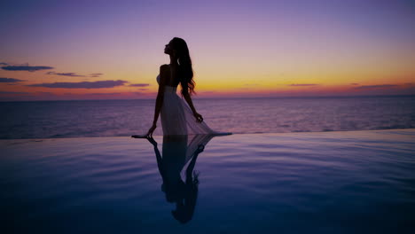 woman in a long dress standing in an infinity pool at sunset