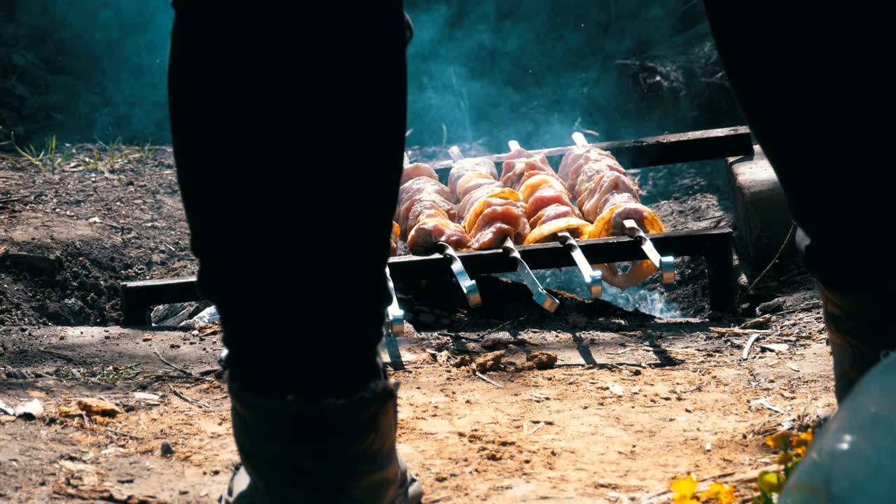 A view of the legs of a man cooking Shish kebab on skewers over a fire in the countryside in the village