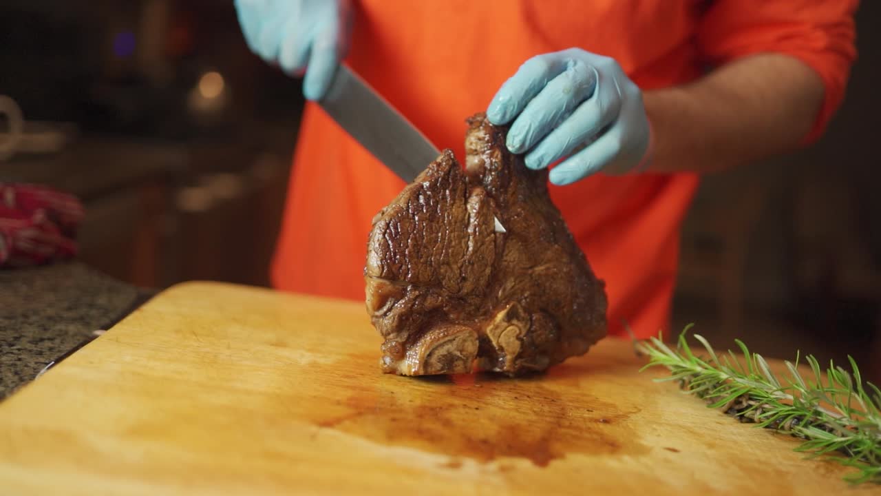 Slo-mo footage of a man wearing gloves cutting a cooked, juicy, steak with a large chef's knife on a wooden cutting board.