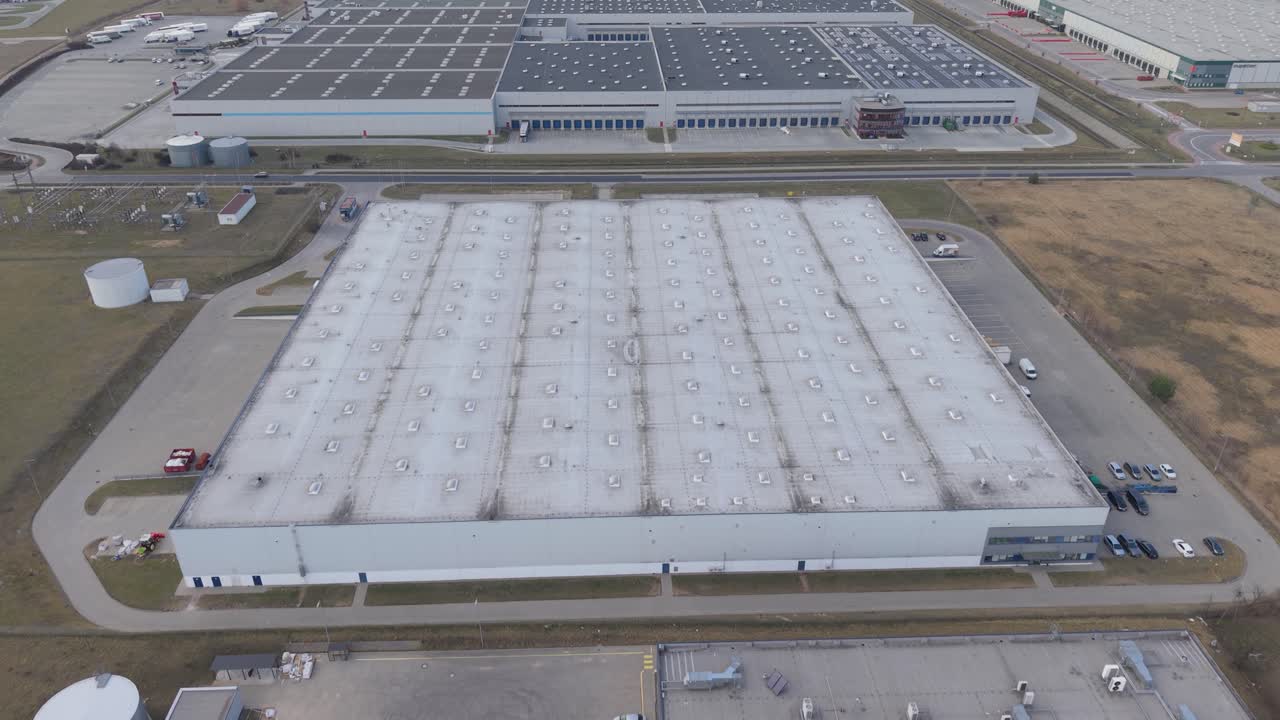 Ampia ripresa aerea con drone di un grande complesso di magazzini logistici e di distribuzione con ampi pannelli solari sul tetto e camion parcheggiati accanto alle banchine di carico. Campi circostanti e strade di accesso visibili.