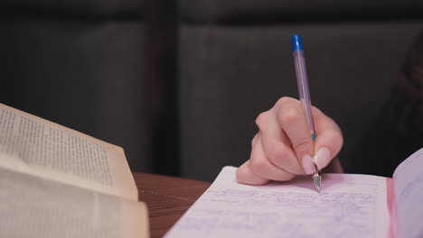 close up view of female hand holding pen and writing in lined pink notebook while aged open book lies on wooden table, capturing intimate academic moment in cozy indoor setting with soft background