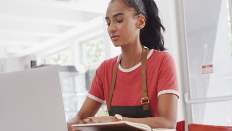 Biracial-female-barista-wearing-apron-using-laptop-at-table-in-her-cafe