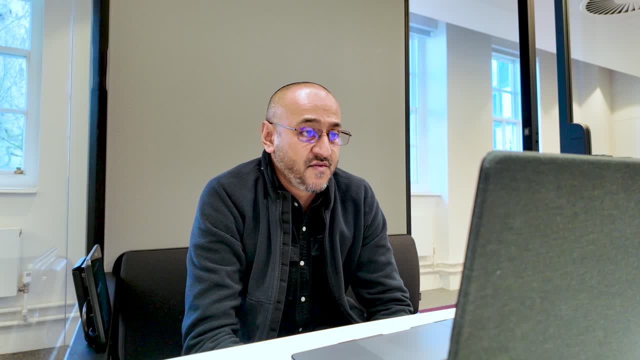 Confident south asian businessman speaking during a webinar in a modern office pod, using a laptop