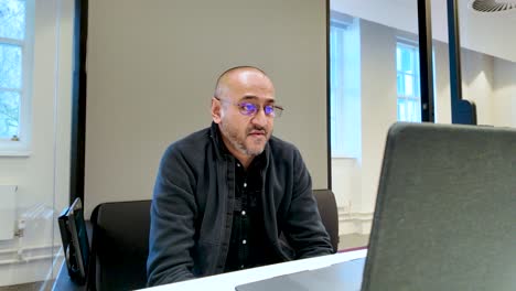 confident south asian businessman speaking during a webinar in a modern office pod, using a laptop