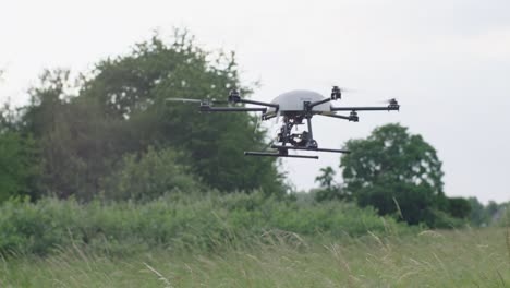 handheld camera captures a black drone with four rotors and a camera, flying over a tall grass field, trees and blue sky in the backdrop