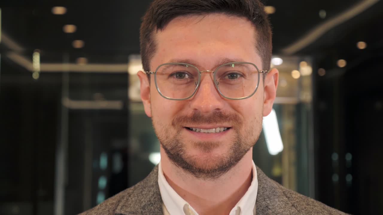 retrato de inteligente barbudo guapo especialista que lleva gafas sonríe encantadoramente mirando a la cámara. rostro positivo de hombre de negocios de pie en la oficina moderna interior. cerca de la expresión facial masculina.
