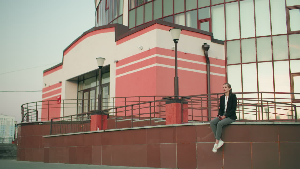 Thoughtful woman seated on fence with legs crossed at ankle holding phone in hands, gazing down in urban setting, surrounded by modern architecture