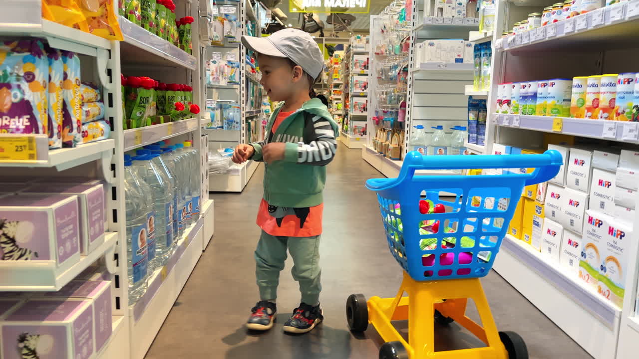 Cheerful baby boy in a cap throws product from a shelf to his cart. Little child shopping in a grocery store.