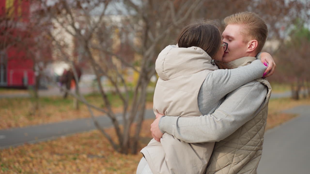 Couple embrace affectionately outdoors as wife in glasses kisses husband with hair fluttering in wind, both in matching jackets, autumn leaves on ground, blurred pedestrians and trees in background