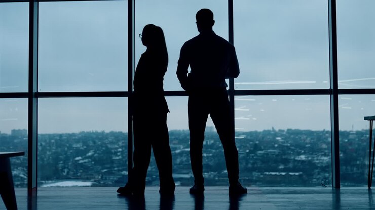 Male silhouette stands his back to camera drinking coffee. Female silhouette walks to and fro speaking on the phone.