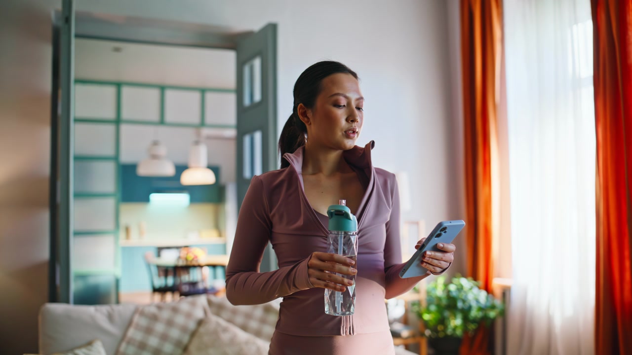 Asian sportswoman holding water bottle browsing phone after home fitness workout