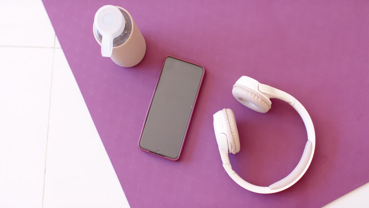 Câmera superior está iniciando a captura, filmando um telefone imóvel, fones de ouvido e uma garrafa de água sobre um tapete roxo para venda.
