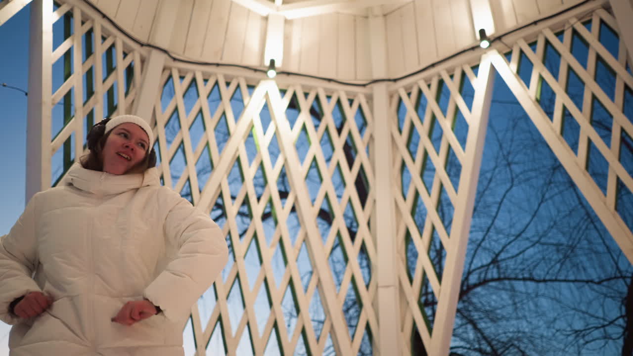 Young artist spinning energetically inside illuminated lattice summer house at dusk wearing white coat beanie and headphones as lights glow around conveying rhythmic movement in evening park setting