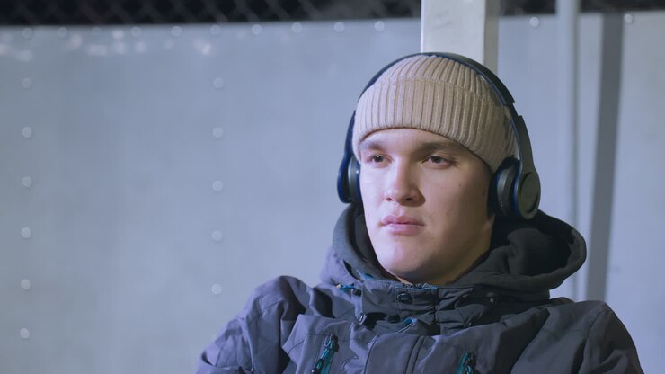 Close up of reflective young man in beanie and headphones, sitting against light background, deep in thought and focused expression, dressed warmly with a subtle sports setting