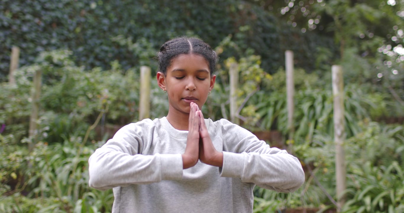 African American teen girl in grey top closing eyes, lowering palms to chest, meditating in garden