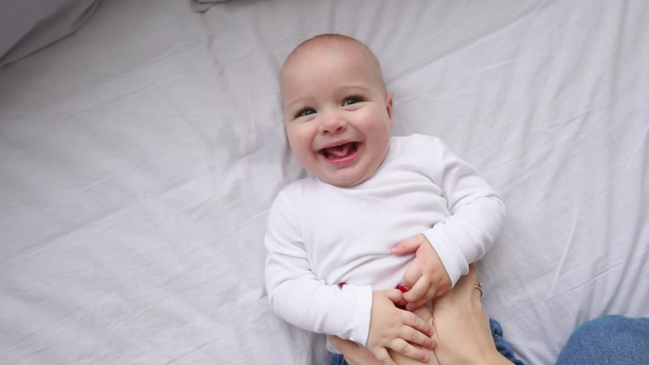 A newborn baby lying on the bed, looking into the camera and around on a white bed sheet, view from above. Infant knows the world, top view