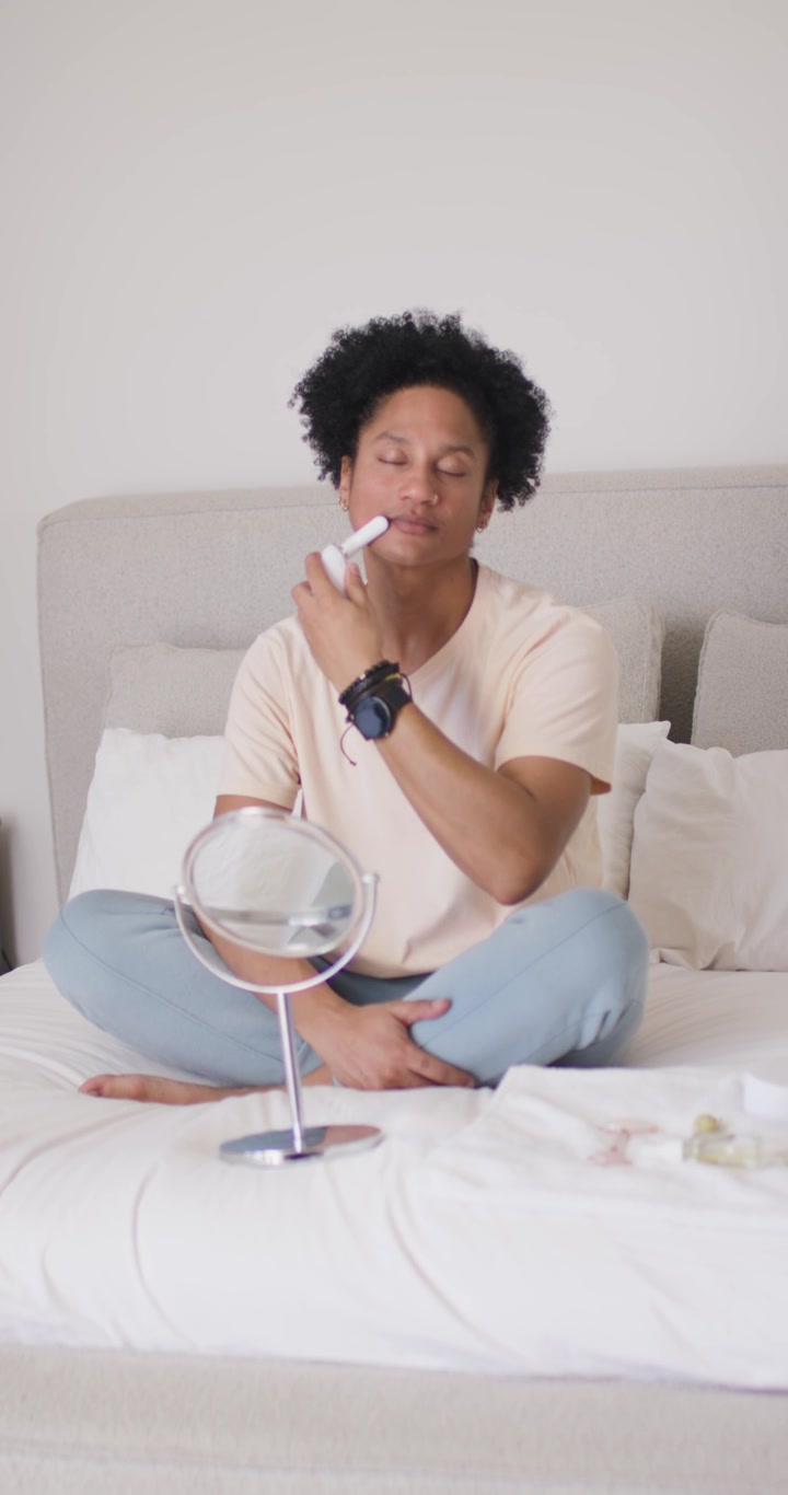 Video verticale: Uomo africano che fa la skincare usando un dispositivo facciale, facendolo scorrere sul viso, sul letto con uno specchio.