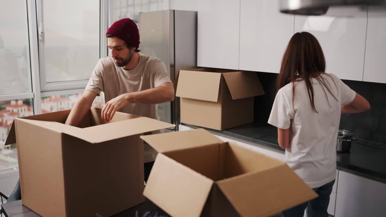 un hombre moreno feliz con un sombrero rojo y una camiseta beige pasando utensilios de cocina a su novia morena con una camisa blanca y sacando cosas de una caja después de mudarse a un nuevo apartamento moderno