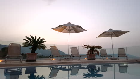 Panning-Left-Shot-of-Reflective-Pool-at-Dusk-with-Sun-Loungers,-Umbrellas---Miniature-Palm-Trees-with-Mountains-in-Distance