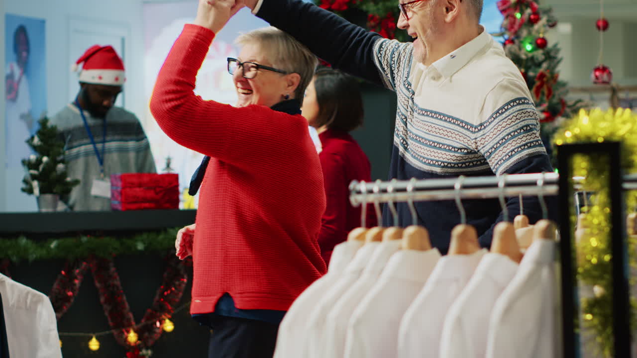 크리스마스 장식 된 옷 가게에서 춤을 추는 즐거운 고령 커플, 가족 구성원들을위한 완벽한 선물을 찾은 후 행복합니다.