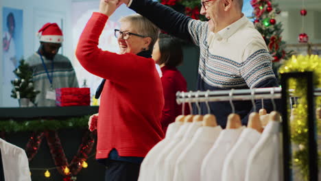 joyful senior couple dancing in christmas decorated clothing store, happy after finding perfect gifts for family members. clients feeling xmas holiday mood spirit while browsing for clothes