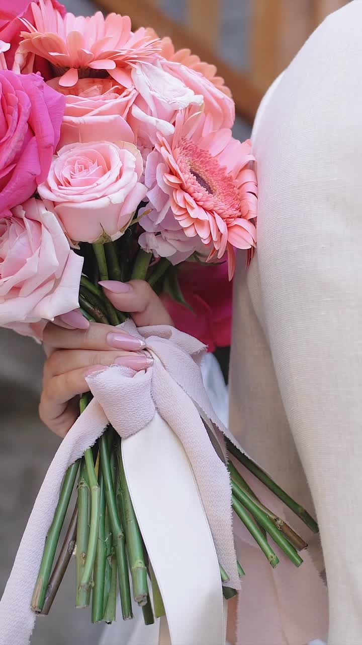 Sposa che tiene un bouquet nuziale rosa e abbraccia lo sposo in abito beige, primo piano verticale al rallentatore