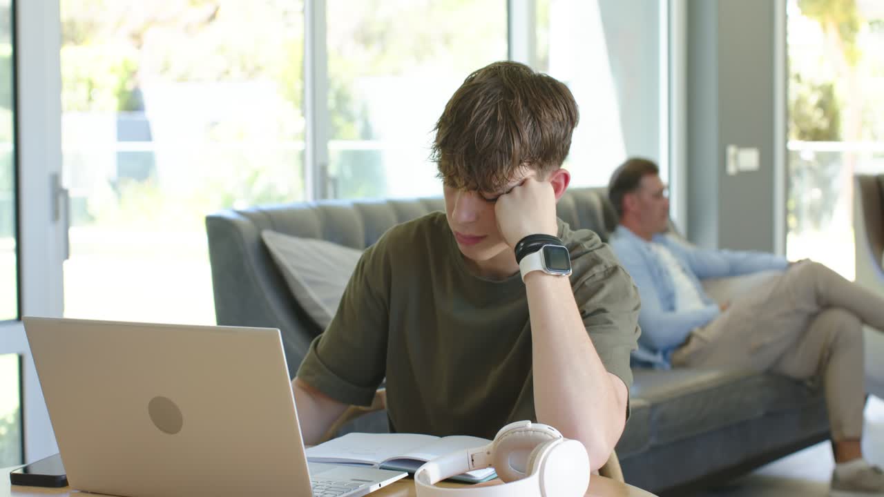 Maschi di casa, un adolescente che fa i compiti a un tavolo rotondo vicino alle finestre, usando un laptop e un quaderno.