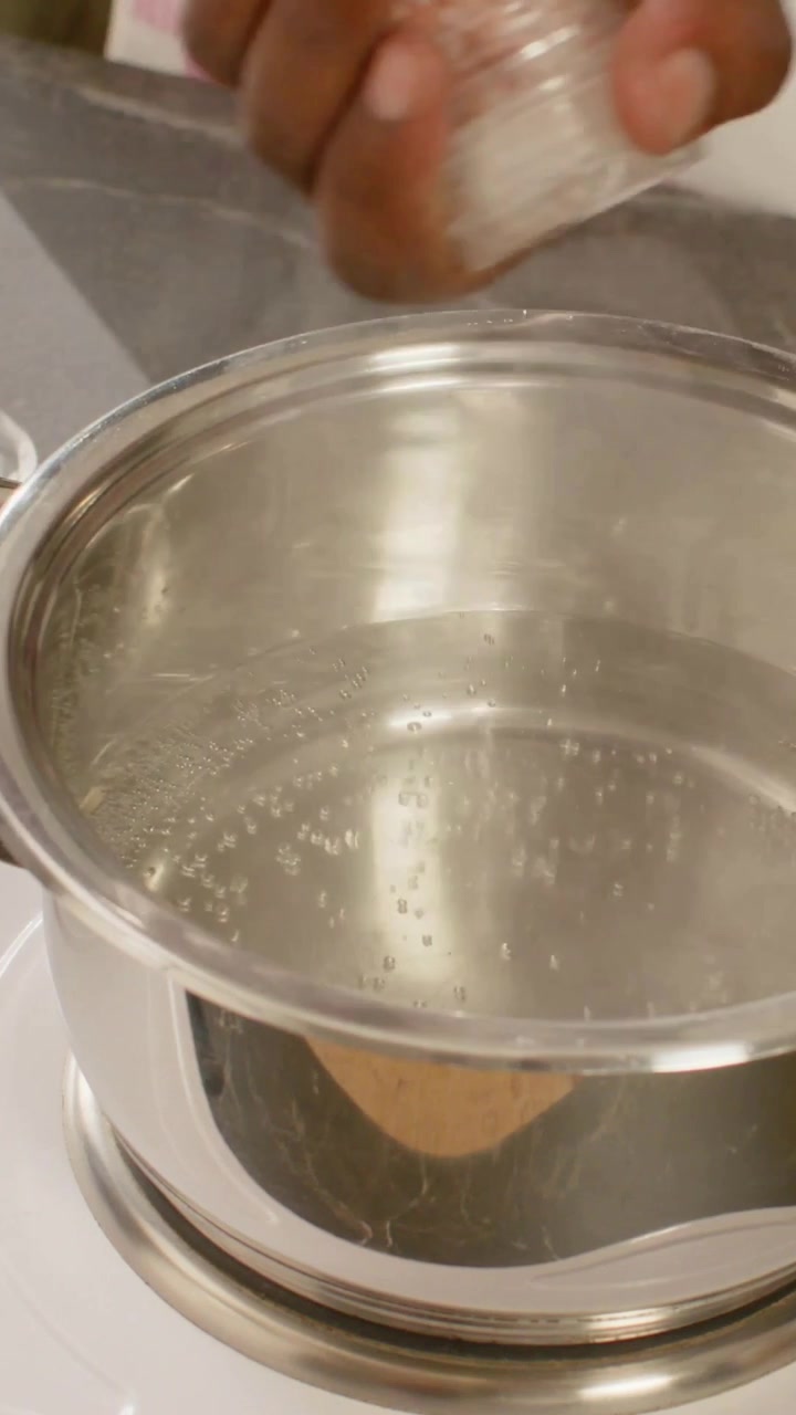 Vertical video: Reaching hand in green top salting pot with jar on hob, lifting pot exposing beads