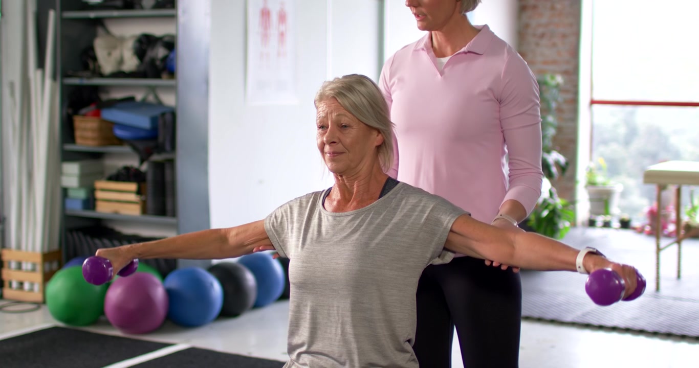 Female coach placing hands shoulders hips guiding grey-clad client lifting weights for rehab in gym