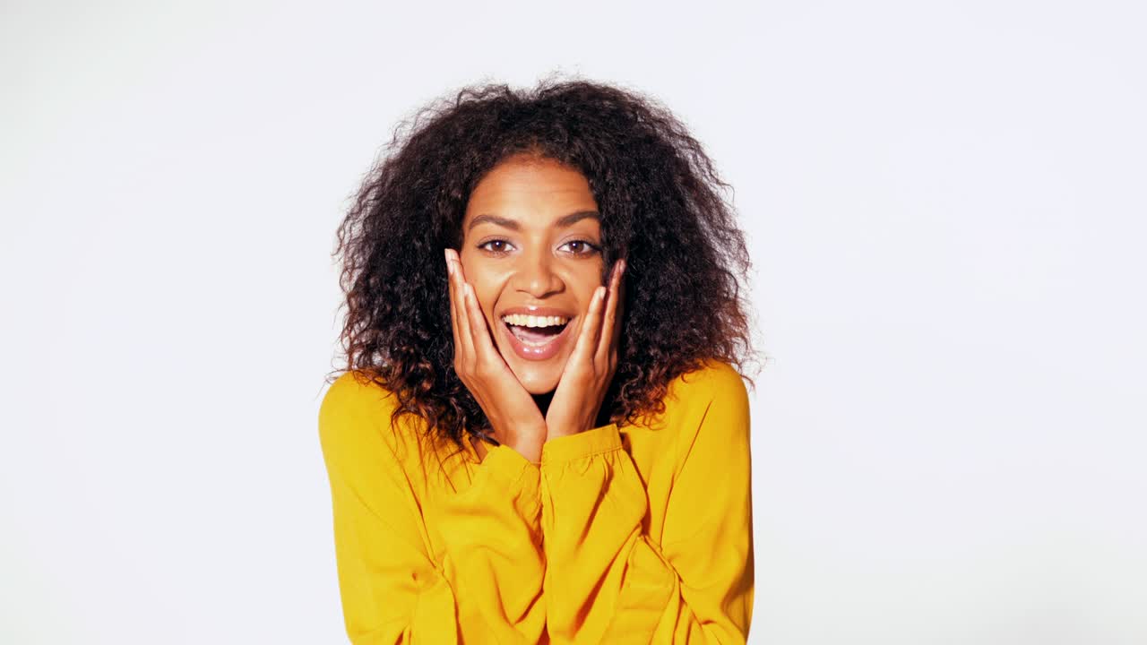 Beautiful african american woman with afro hair in yellow wear smiling, pleasantly surprised to camera over white wall background. Cute mixed race girl's portrait with amazement