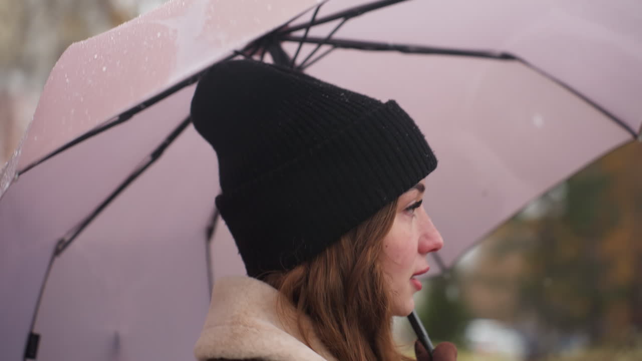 Close shot of happy lady holding umbrella as wind blows gently with light snowfall running outdoors wearing black knit cap and brown shearling jacket strands of hair blowing creating cozy winter vibe