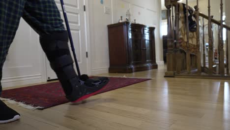 hombre con una lesión en el tobillo usa una bota ortopédica y un bastón para caminar por su casa