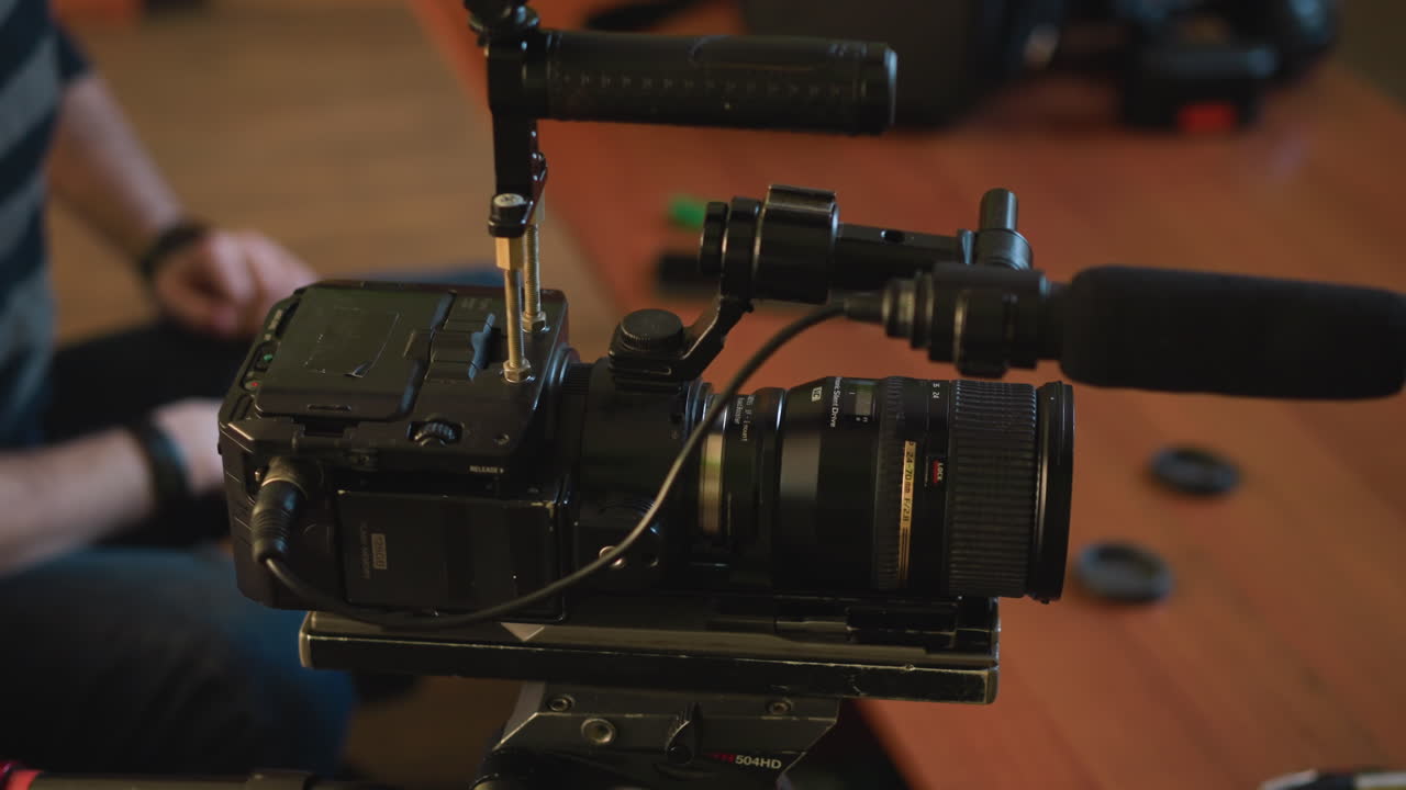 Close-up of camera tripod quick release plate on wooden table. Two lens caps placed nearby. Background includes couch and soft indoor lighting creating a relaxed environment