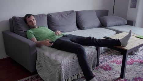 skinny young man with dark hair and green t-shirt switching over the channels on remote control sitting on the couch in relaxed position watching tv with his foot on the wooden table