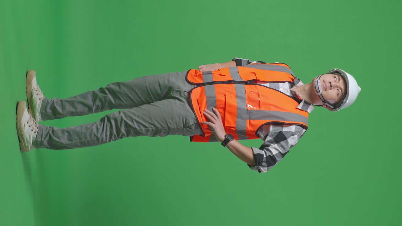 Full Body Side View Of Asian Male Engineer Wearing Safety Helmet Looking Around While Standing With Arms Akimbo In The Green Screen Background Studio