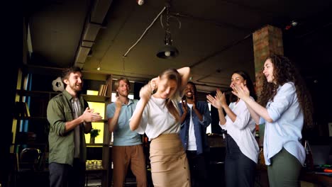 company employees are dancing at corporate party in modern loft style office, attractive young men and women are having fun, clapping hands and laughing.