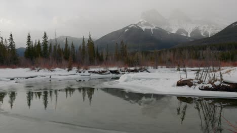 Mañana-Brumosa-En-Canmore,-Alberta,-Canadá,-Con-Las-Icónicas-Montañas-De-Las-Tres-Hermanas-Envueltas-En-Nieve,-Mientras-Un-Estanque-Tranquilo-Adorna-El-Primer-Plano,-Ofreciendo-Una-Visión-De-La-Belleza-Etérea-De-La-Naturaleza.