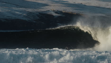 surfer carves and throws spray glistening in light with snow in background of heavy wave