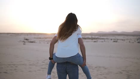 Happy-and-cute-adorable-adult-couple-in-white-shirts-and-jeans-man-with-woman-girlfriend-on-piggy-back-running-by-desert-crazy-in-love,-emotions-and-relationship.-Whirling-around.-Mild-sunset-on-background